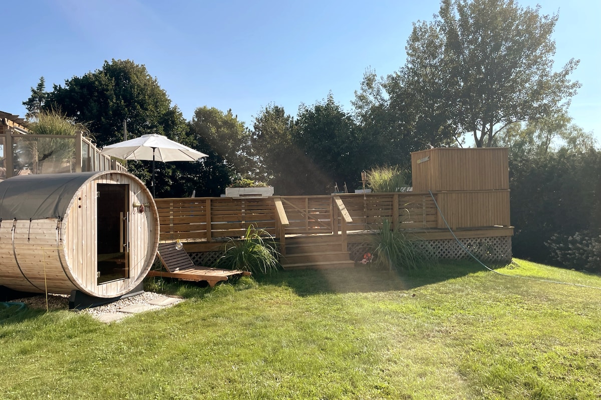 A cedar barrel sauna is situated on a grassy area, accompanied by a wooden deck. An outdoor umbrella provides shade near the seating area. Lush greenery surrounds the space, while sunlight casts a warm glow over the setting.