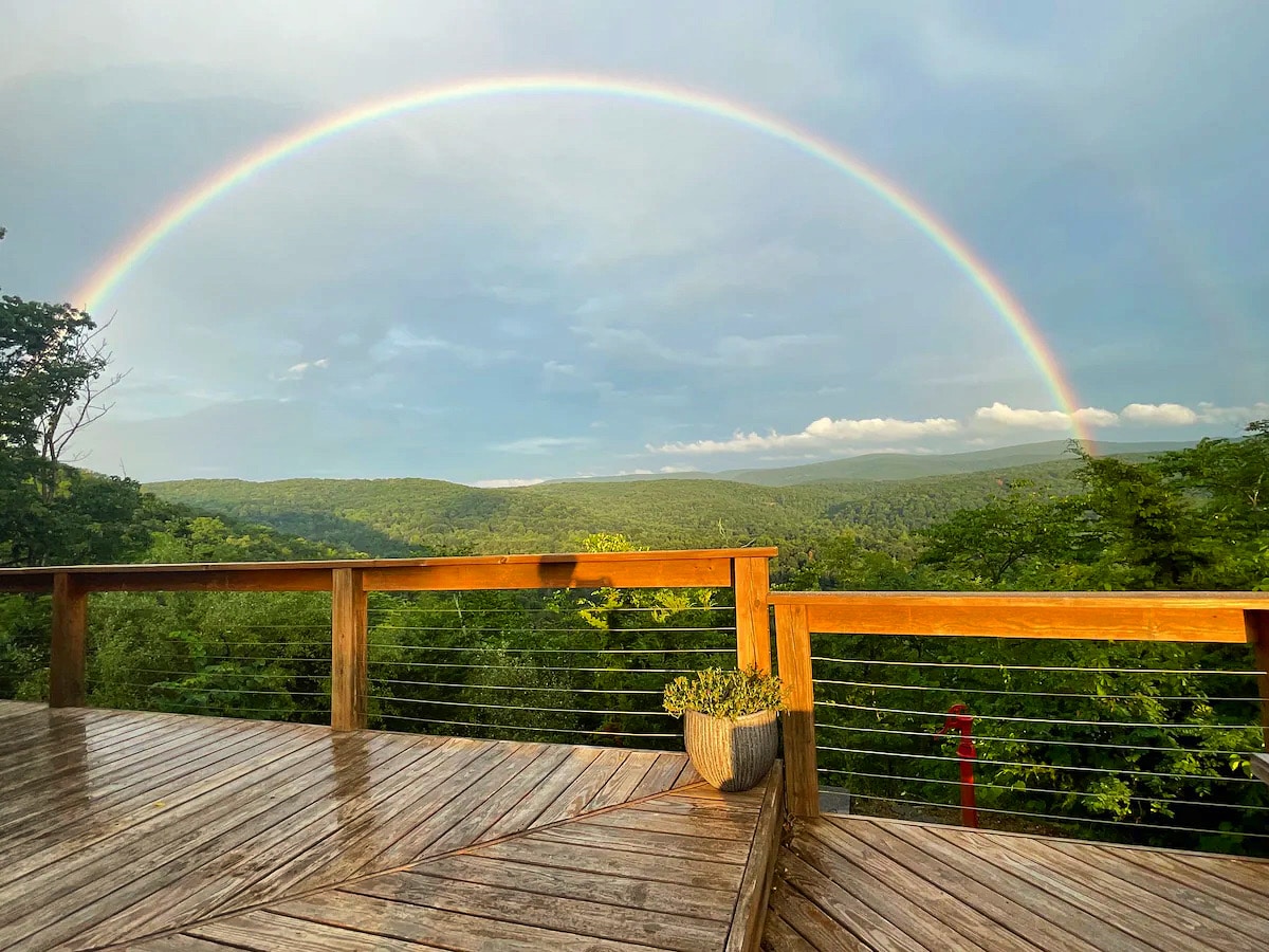 A spacious wooden deck is displayed, featuring a vibrant rainbow arching across a panoramic view of lush green mountains and valleys. A planter with greenery sits on the deck, surrounded by the serene landscape after a light rain.