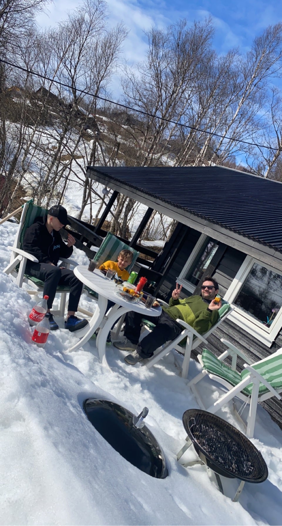 An outdoor table is set on a snowy surface, featuring a variety of colorful food items and drinks. Two figures are seated on chairs, enjoying a meal. The cozy cabin is visible in the background, surrounded by trees and clear skies.
