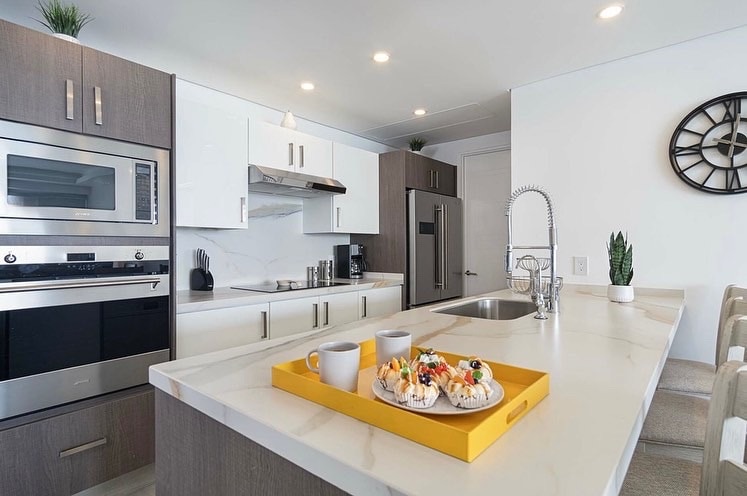 A modern kitchen showcases sleek cabinetry and stainless steel appliances, complemented by a spacious island. A tray with colorful breakfast items and two mugs rests on the countertop, while natural light illuminates the area from overhead fixtures.