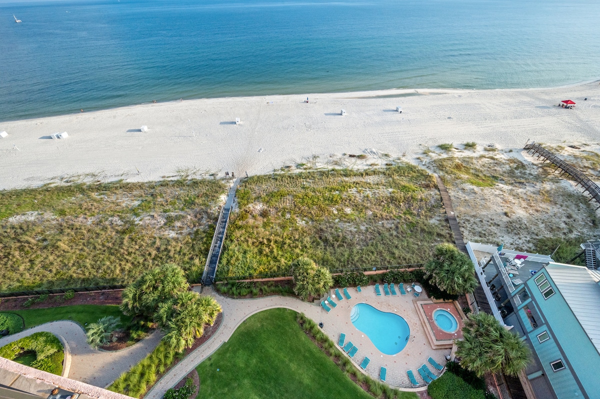An aerial view captures a sandy beach stretching along the coastline, with beach chairs arranged on the shore. Below, a landscaped area features a pool surrounded by lounge chairs and a hot tub, complemented by lush greenery and pathways leading to the beach.