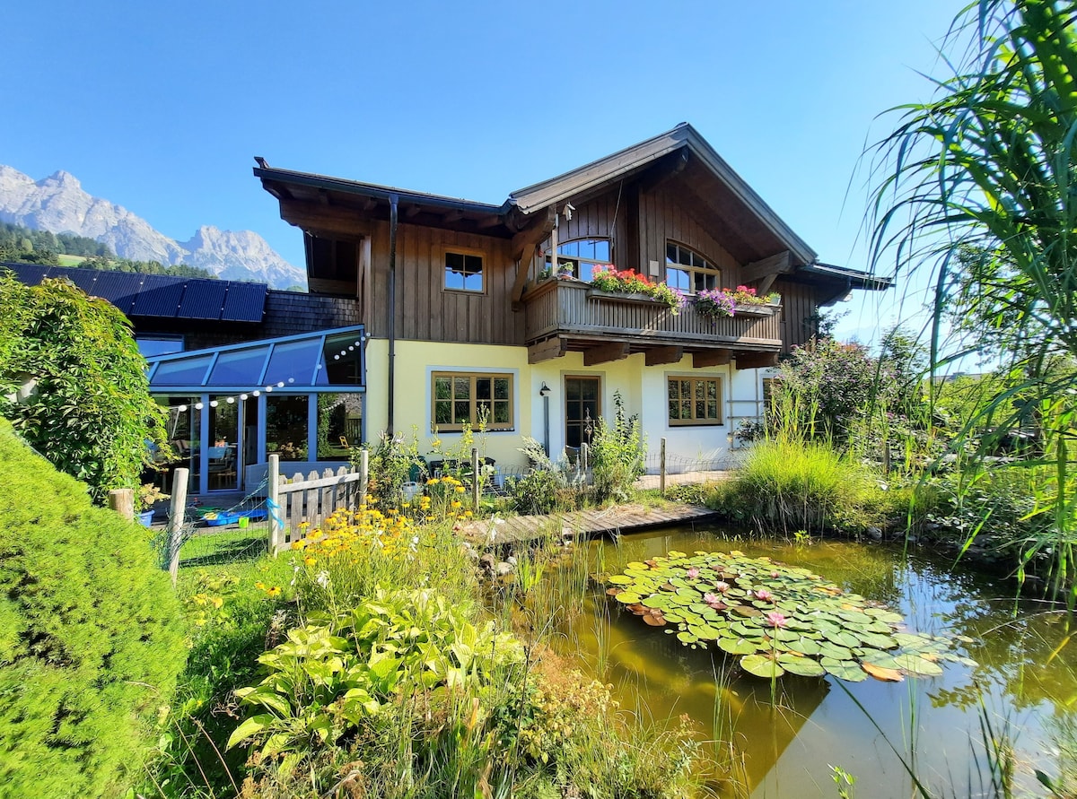 The charming two-story house is surrounded by lush greenery and features a serene pond with lily pads in the foreground. Colorful flowers adorn the balcony and garden, complementing the natural landscape with mountains visible in the background under a clear blue sky.