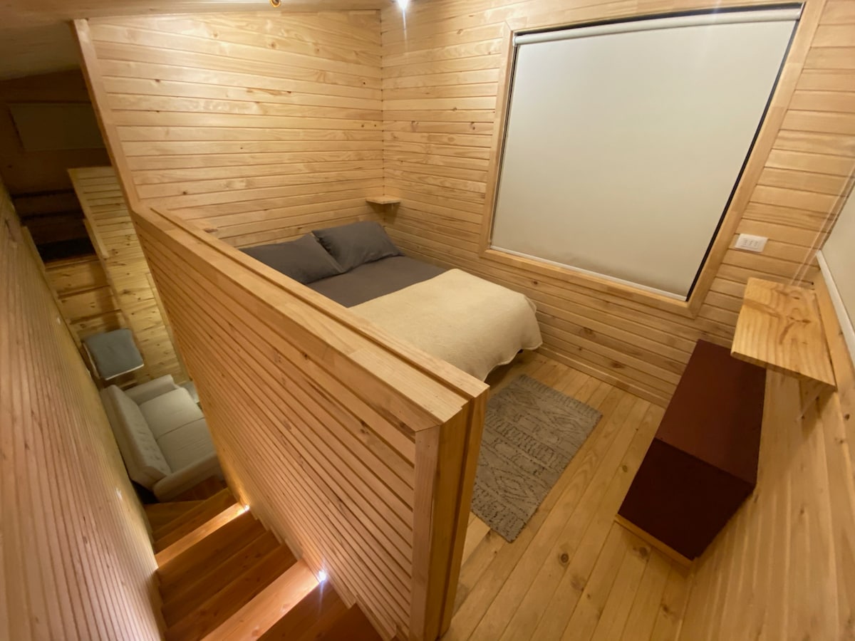 A cozy sleeping area features a spacious bed covered with soft linens, positioned against wooden planked walls. Natural light enters through a large window, which is framed by simple, light-colored shades. A wooden side table and a textured rug complement the warm ambiance.