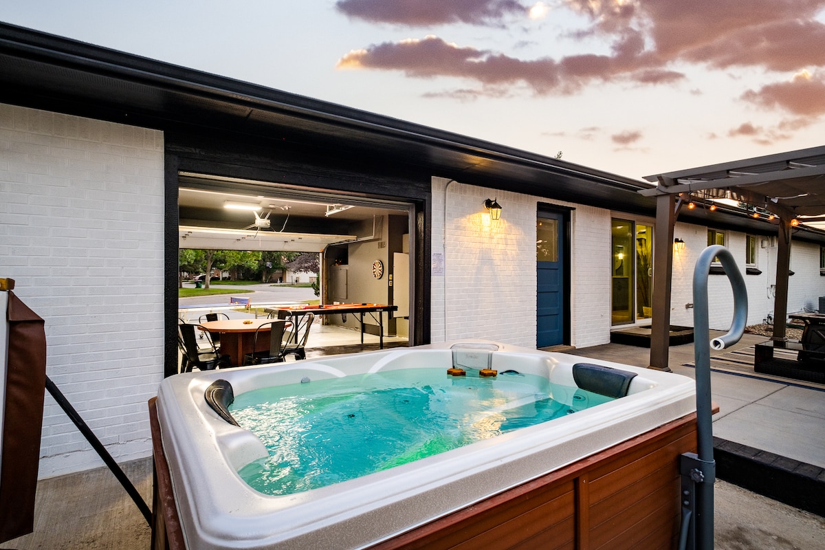 A newly installed hot tub sits outside, surrounded by a wooden deck. Nearby, an outdoor game area with a pool table and barbecue grill is visible, under a covered lounge space. The scene is complemented by a soft twilight sky.