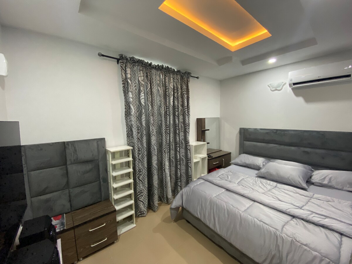 A contemporary bedroom features a spacious bed with layered gray bedding. A dark curtain frames the window, and the room is equipped with a sleek dresser and a white shelving unit. Soft lighting highlights the textured ceiling, creating a calming ambiance.