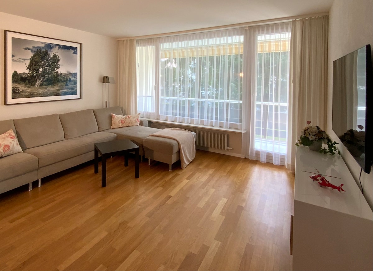 A comfortable living area features a spacious gray sectional sofa and a dark wood coffee table, with a light-colored throw draped over the arm. Large windows allow natural light to fill the room, complemented by soft curtains. A television is mounted on a sleek white console.