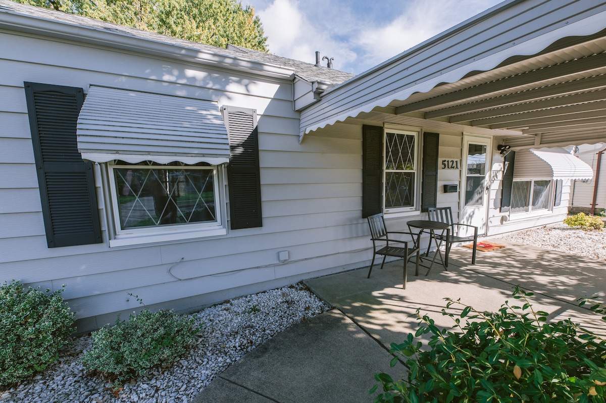 An inviting covered porch features a small table and two chairs, set against a backdrop of light-colored siding and neatly trimmed shrubbery. Windows adorned with classic shutters complete the charming exterior, while a concrete pathway leads to the front door.