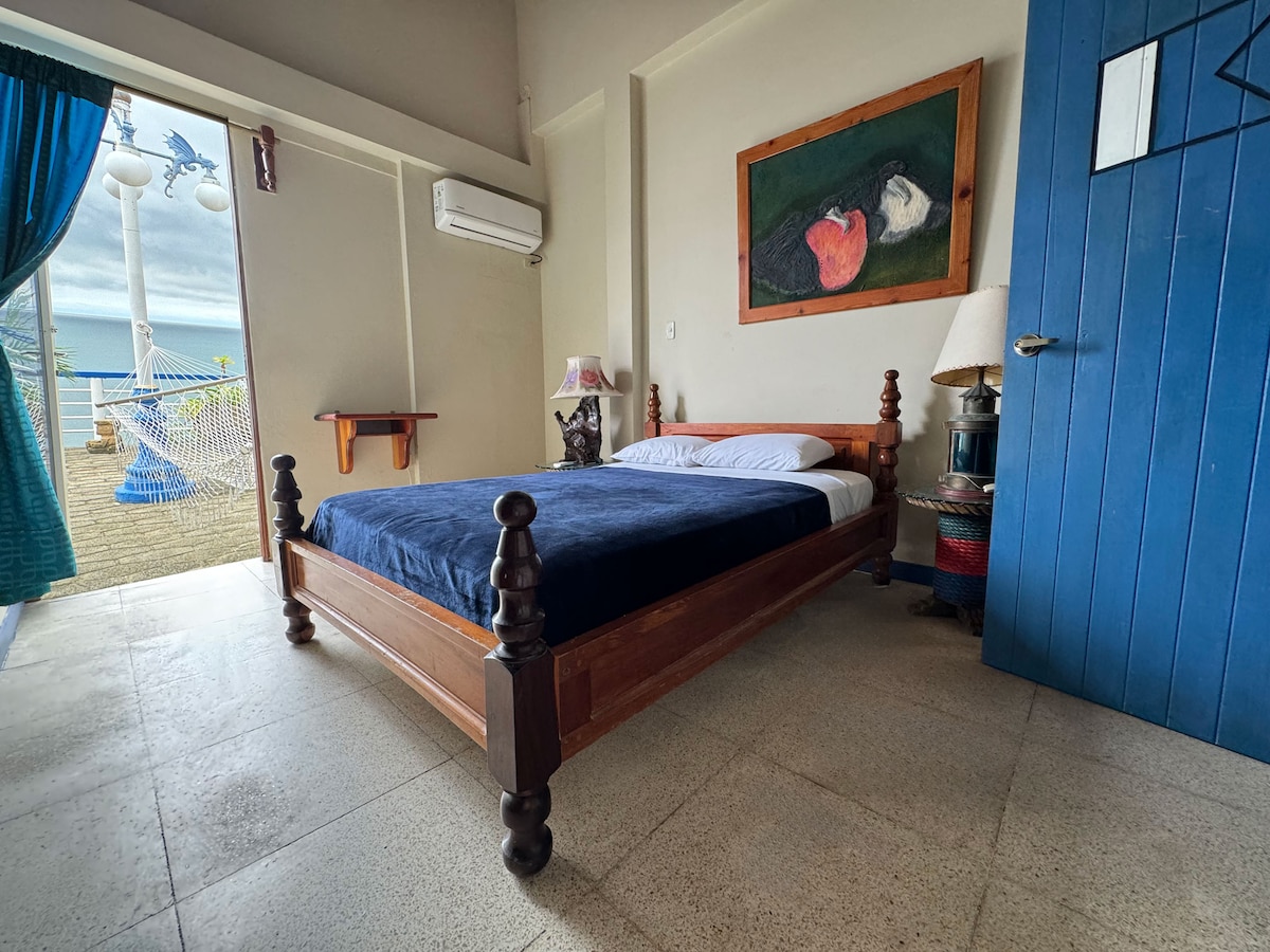 A spacious bedroom is equipped with a wooden bed featuring a dark blue coverlet. A large window offers access to a private balcony with ocean views. An air conditioning unit and artistic decor enhance the space. Natural light brightens the room through the open door.