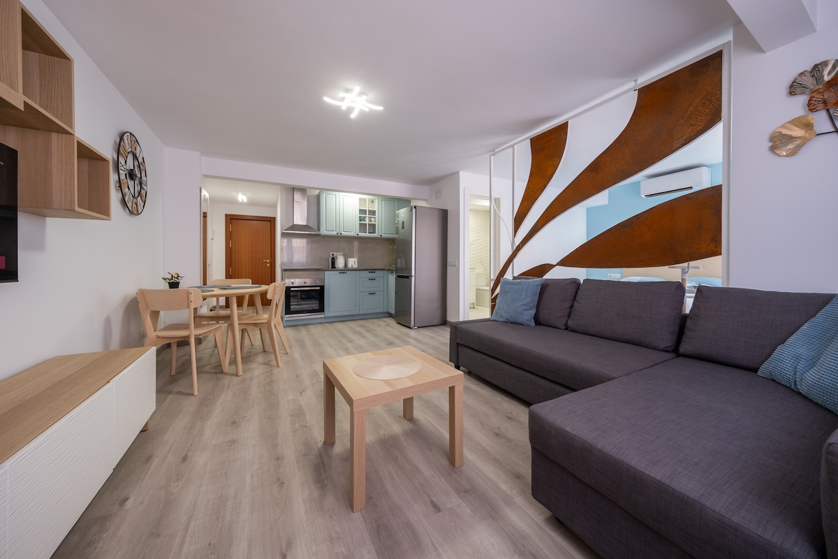 A modern studio layout is shown, featuring a spacious living area with a comfortable L-shaped sofa and a wooden coffee table. The open kitchen is equipped with sleek cabinetry and appliances, while a dining table with wooden chairs is present in the background.