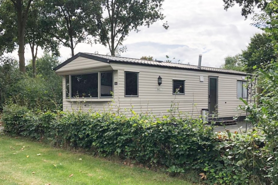 A light-colored caravan is surrounded by greenery, situated on a spacious campsite. Large windows provide ample natural light, while a sloping roof contributes to a classic aesthetic. The exterior is bordered by a neatly trimmed hedge, enhancing the connection with nature.
