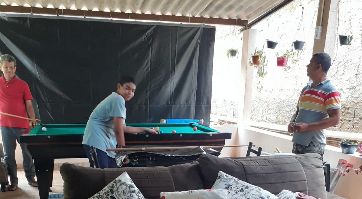 A billiards table is positioned under a covered outdoor space, surrounded by potted plants along the walls. Two individuals are engaged in a game, while a comfortable seating area is visible in the foreground, featuring a brown couch adorned with decorative pillows.