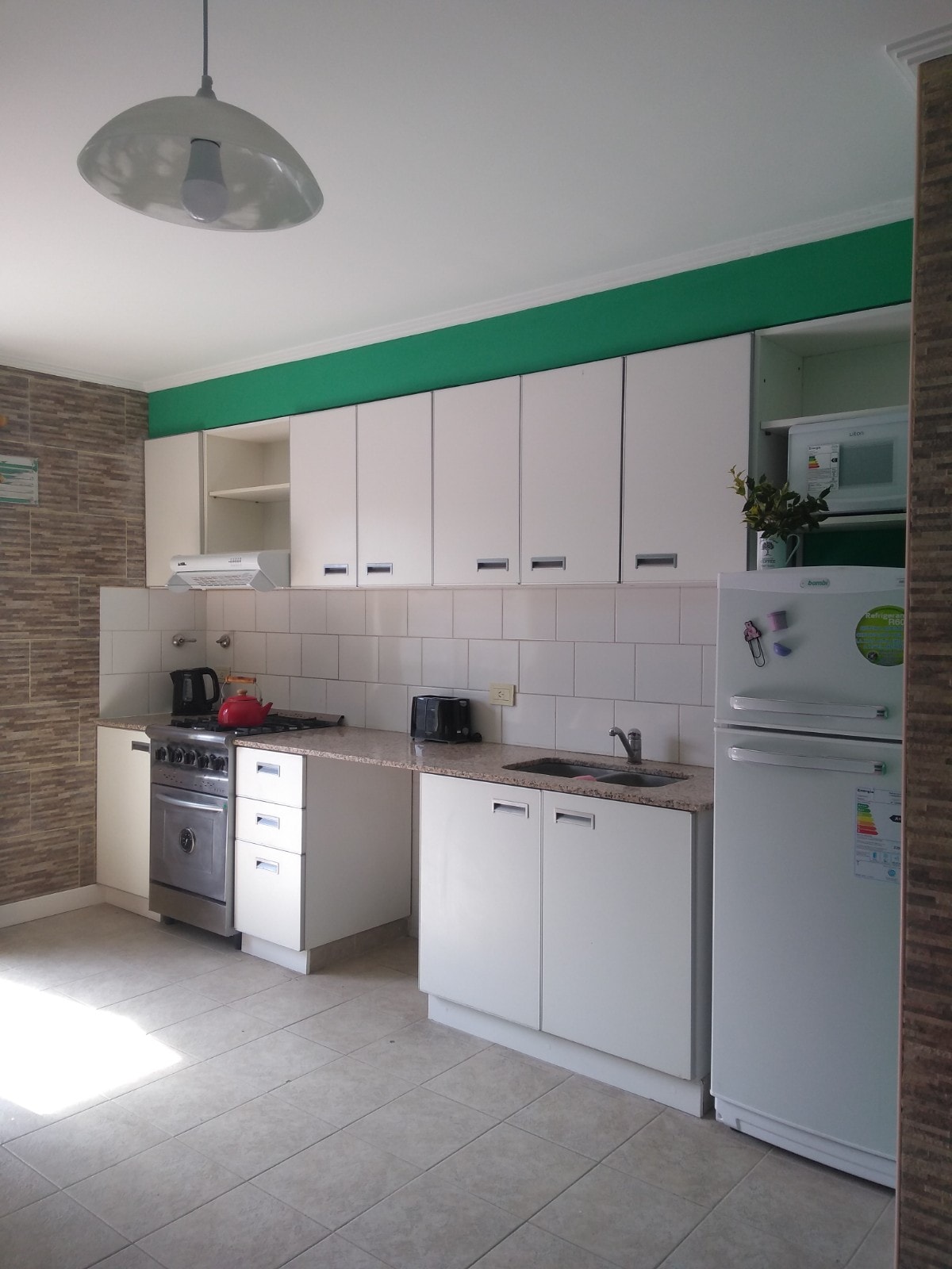 A modern kitchen space features a combination of white cabinets and green accents. It includes a stainless steel stove, a refrigerator, and various small appliances. Countertops are equipped with essential prep areas, while upper cabinets provide storage. Natural light brightens the room through a nearby window.