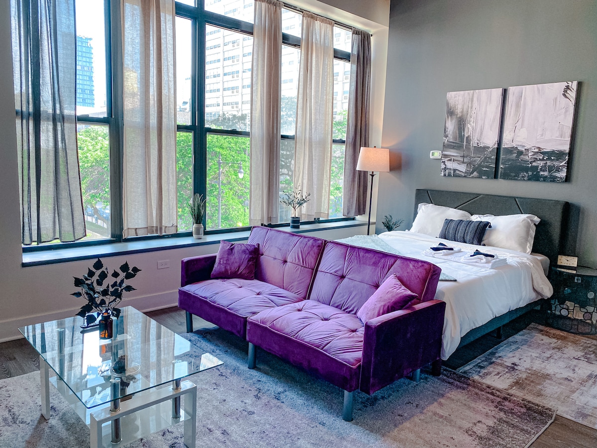 A spacious room features a queen-size bed with crisp white bedding. A plush purple sofa sits in front of large windows, dressed with sheer curtains that allow natural light to filter in. A glass coffee table rests on a soft area rug, enhancing the modern decor.