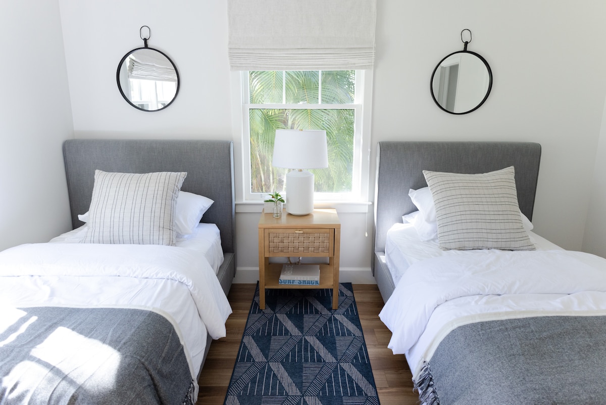 The bright bedroom features two spacious beds with simple gray headboards, oriented parallel to a light-colored wooden nightstand. A small table lamp and a decorative plant sit atop the nightstand, while large windows allow natural light to illuminate the space.