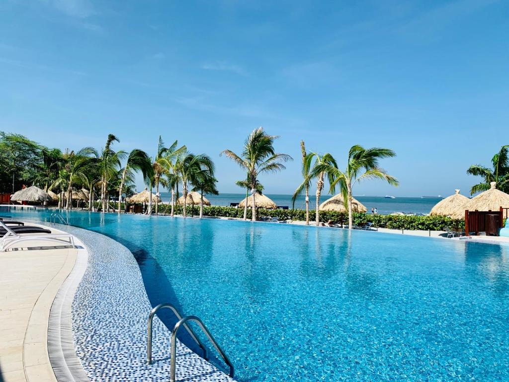 A large swimming pool reflects the clear blue sky, surrounded by palm trees gently swaying in the breeze. Thatched-roof cabanas are positioned along the shoreline, offering shaded spots near the water's edge. The pool area features a smooth edge, inviting relaxation and enjoyment.