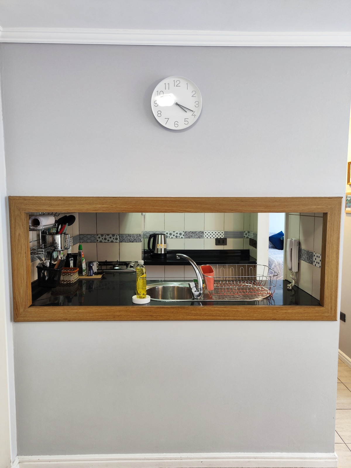 A well-designed kitchen area is visible through a rectangular wooden frame. A clock is mounted on the wall above, indicating the time. The countertop features a sink and various kitchen items, including a drying rack. The space is organized and functional.