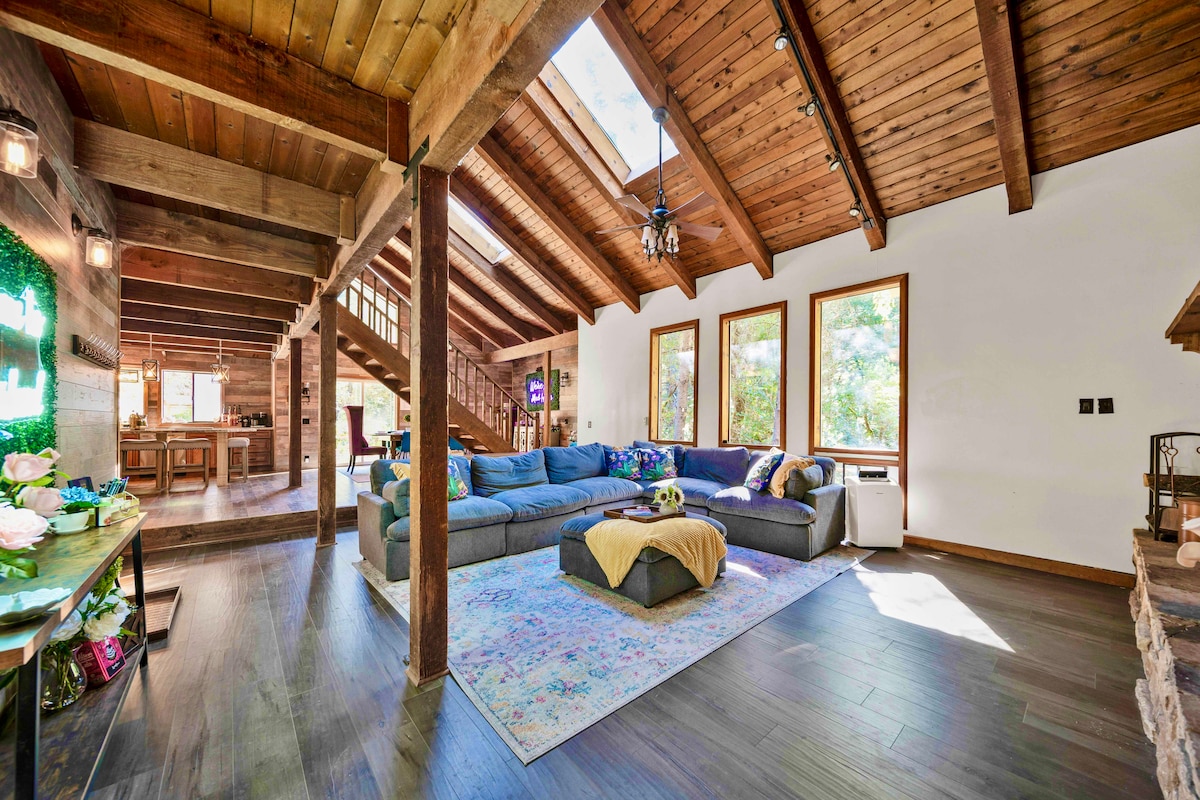 The spacious living area features vaulted wooden ceilings and large windows allowing natural light to illuminate the space. A plush sectional couch, adorned with colorful throws, invites relaxation. In the background, a glimpse of the kitchen is visible, showcasing an open design that connects both spaces.