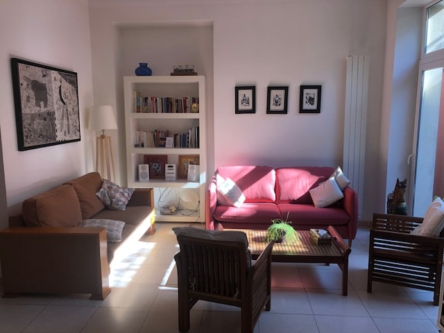A bright and inviting living area is furnished with a combination of sofas and armchairs. A bookshelf filled with books is positioned against the wall, while framed black-and-white photographs hang above. Natural light filters in through large windows, creating a comfortable atmosphere.