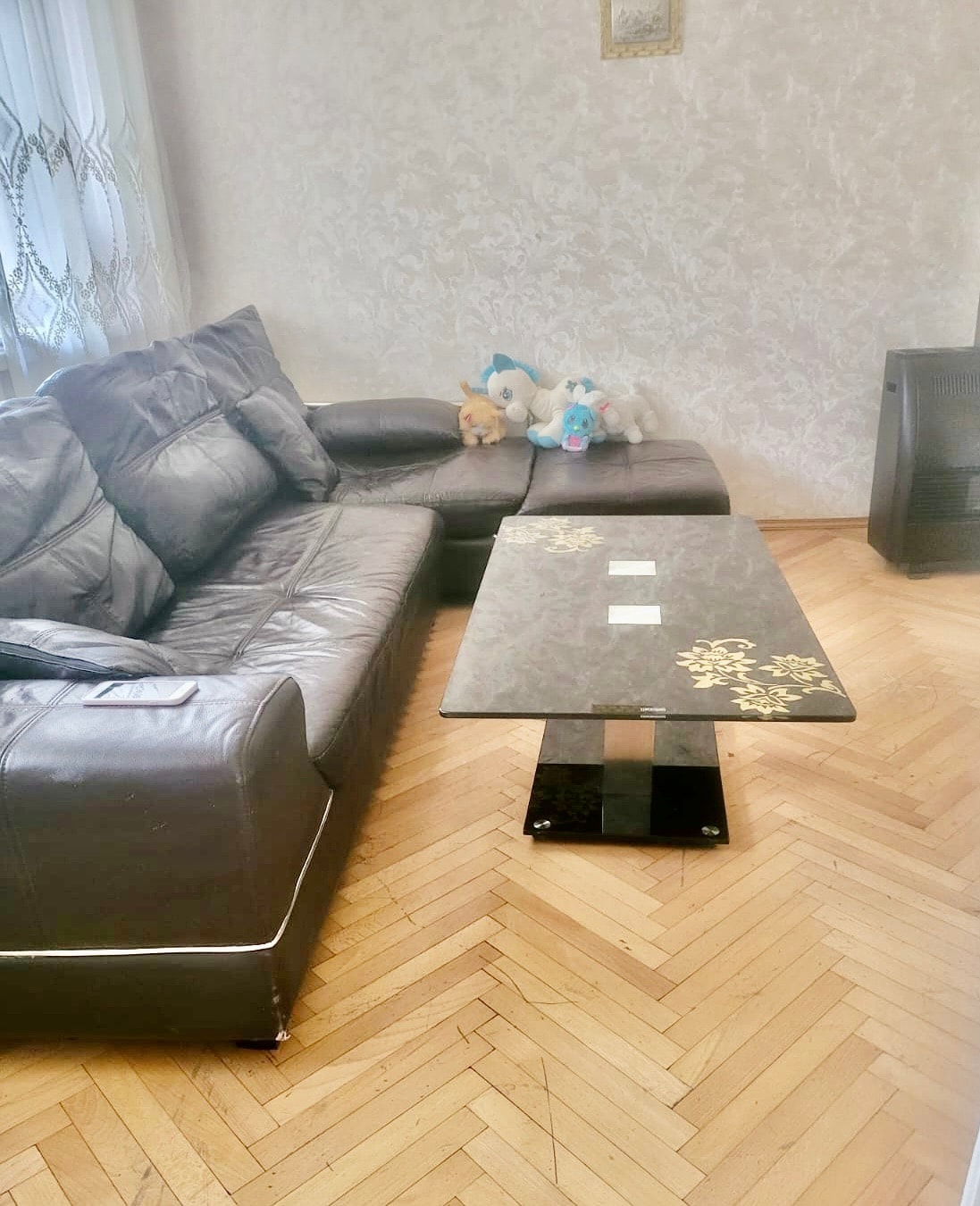 A spacious seating area is highlighted by a large brown leather sectional couch and a sleek coffee table with floral accents. Plush toys are displayed on the couch, adding a touch of charm to the inviting space. Warm wood flooring complements the light-colored walls.