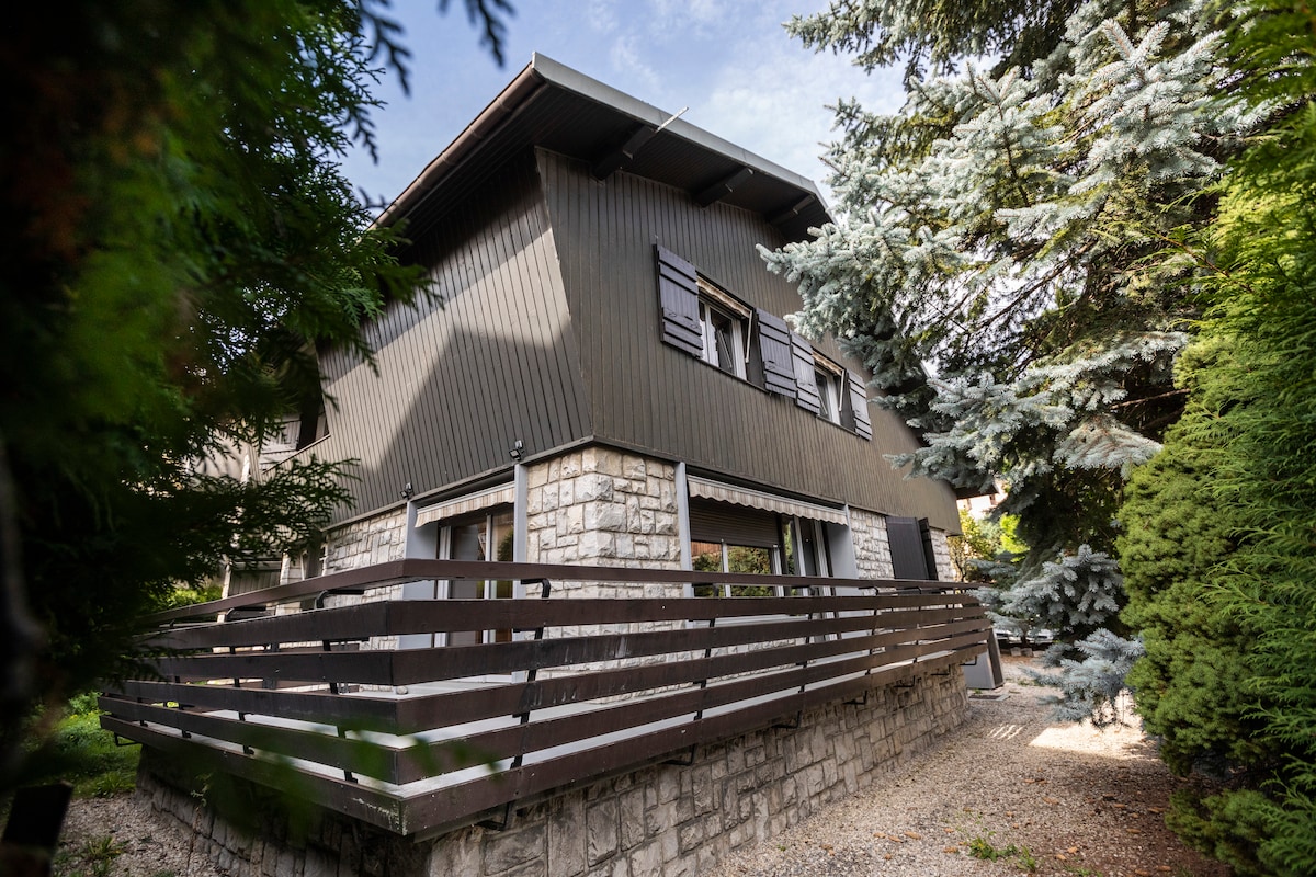 A two-story family residence is framed by lush greenery, featuring a mix of wooden and stone exteriors. Large windows are visible, allowing natural light to enter. A shaded deck with wooden railing provides an outdoor lounging space amidst the surrounding trees.