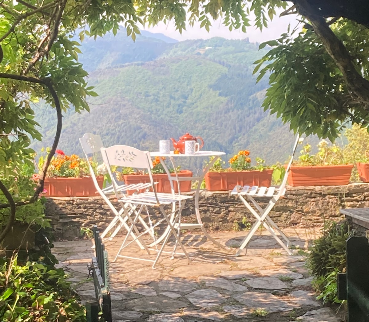 A charming outdoor terrace is set among stone walls, featuring a round table flanked by white chairs. Flower pots add vibrant colors, while a scenic view of the surrounding mountains is visible in the background, enhancing the peaceful ambiance.
