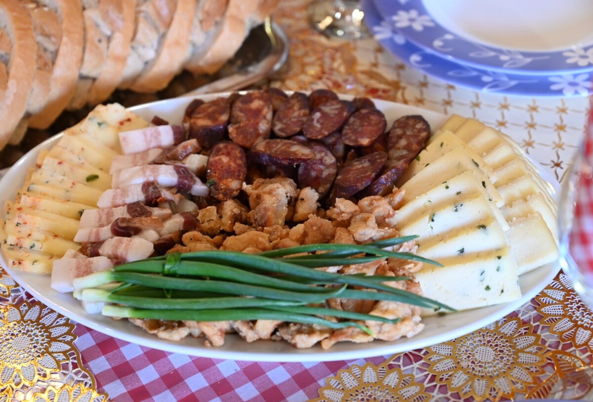 A large platter is arranged with a variety of cured meats, including sliced sausages and smoked bacon, paired with an assortment of cheeses. Fresh green onions are placed across the center, offering a vibrant touch. In the background, loaves of bread are visible.