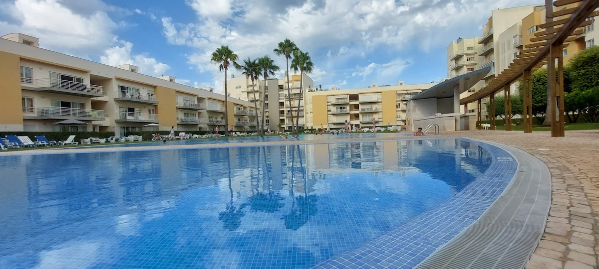 A large outdoor swimming pool is depicted, with clear blue water reflecting the cloudy sky. Surrounding the pool are palm trees and lounge chairs. The buildings of the condominium are visible in the background, creating a spacious and inviting environment.