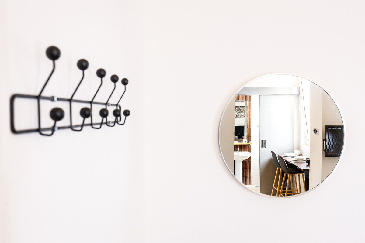 A black coat rack with multiple hooks is mounted on a light-colored wall. A round mirror reflects a glimpse of the dining area, featuring wooden stools arranged around a table, contributing to the functional and stylish ambiance.