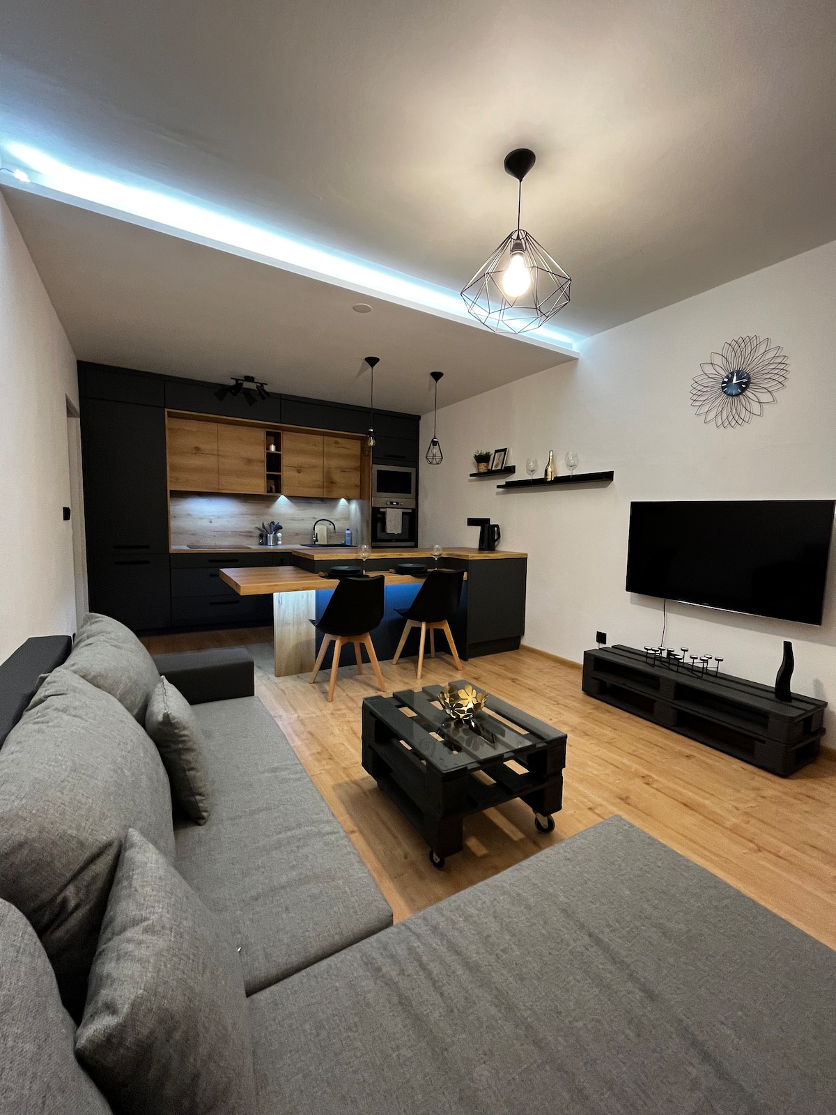 A modern living area features a gray sofa and a stylish coffee table, with a kitchen and dining space in the background. The sleek cabinetry combines dark tones with natural wood accents. A wall-mounted television is visible, along with decorative items on shelves.