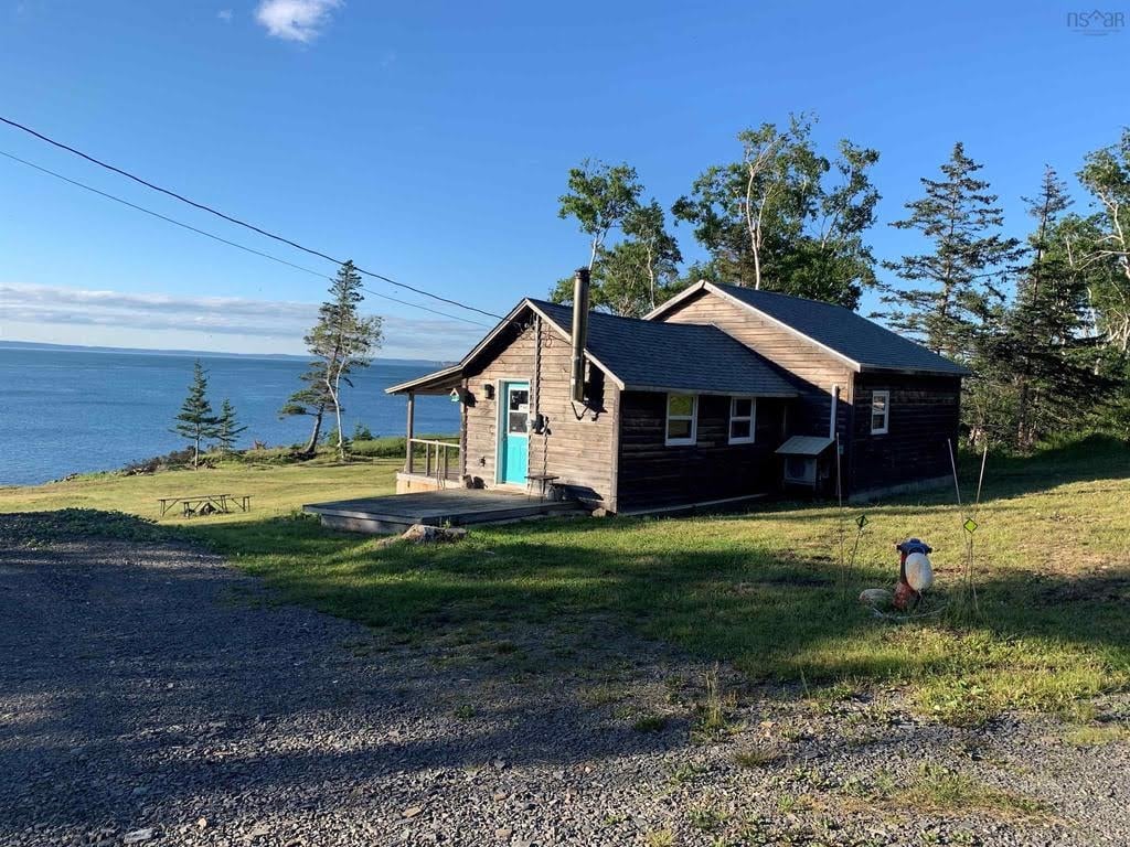 A charming cottage is set against a backdrop of open water and trees. The exterior features a light-colored wood finish, with a blue front door adding a pop of color. The patio area is visible, providing outdoor relaxation space with sweeping views of the landscape.