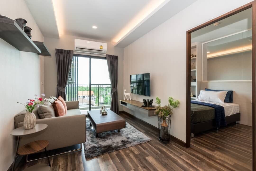 A modern living area features a comfortable sofa paired with decorative pillows, a coffee table, and a large wall-mounted television. Natural light enters through sliding glass doors, leading to a balcony. A cozy sleeping area is visible in the background, separated by a partial wall.