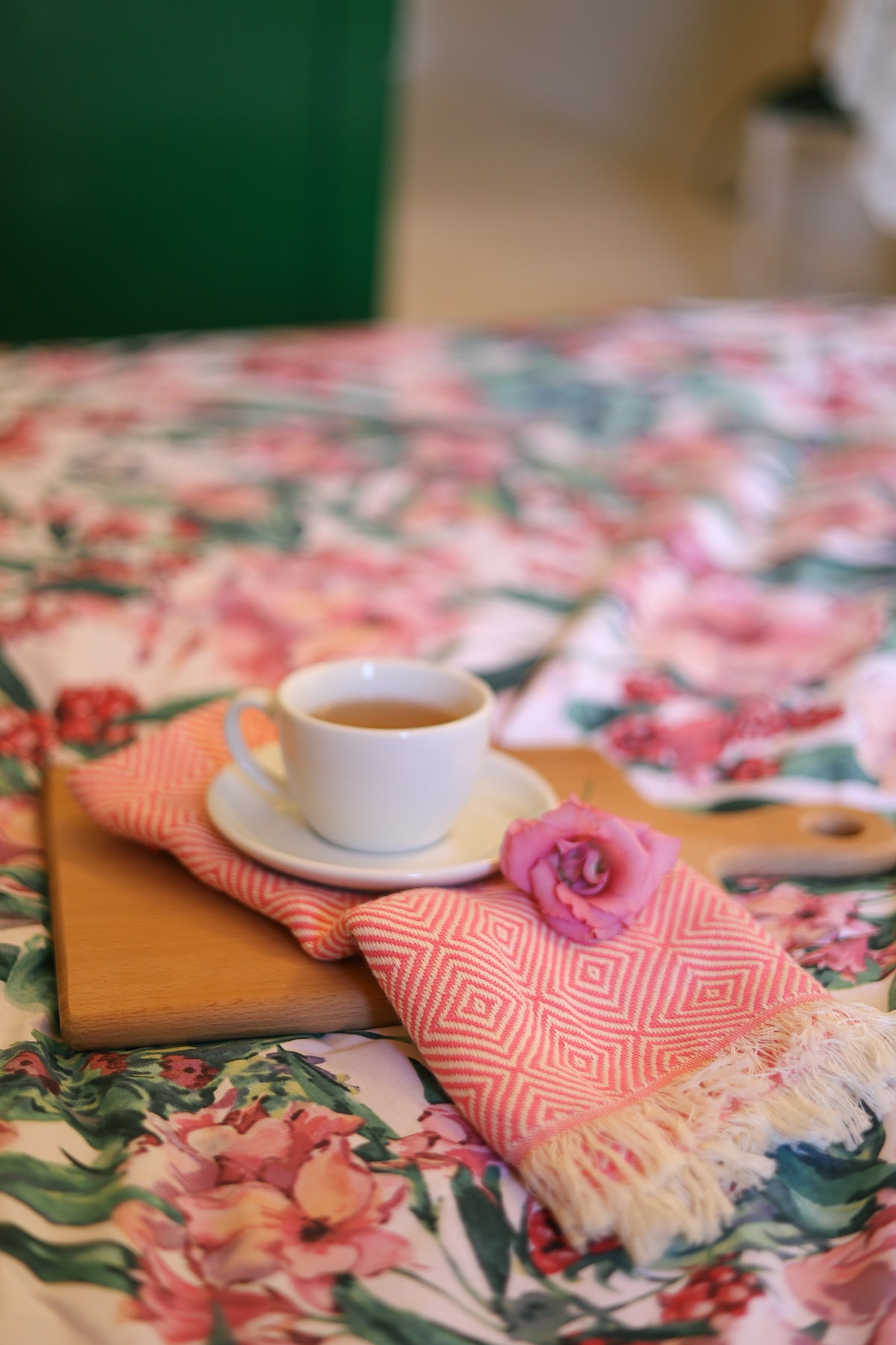 A wooden tray rests on a floral-patterned bedspread, featuring vibrant pink flowers. A white cup of tea sits atop the tray, accompanied by a pink floral napkin and a delicate rose, creating a serene and inviting scene.