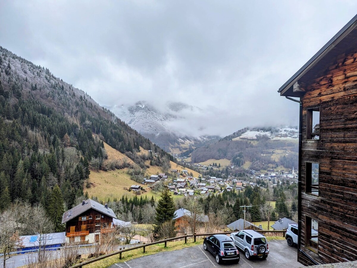 A panoramic view of rolling hills and valleys surrounded by mountains is captured, with a mix of greenery and patches of snow. Wooden chalets dot the landscape below, while low-hanging clouds add a touch of softness to the scene.