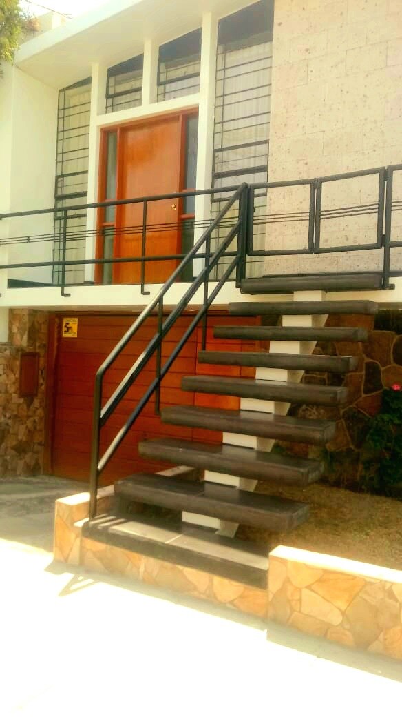 An exterior view of a multi-level building is presented, featuring a wooden front door and an ascending concrete staircase with a metal railing. Stone wall elements and a manicured pathway add to the architectural appeal, with natural landscaping visible in the surroundings.