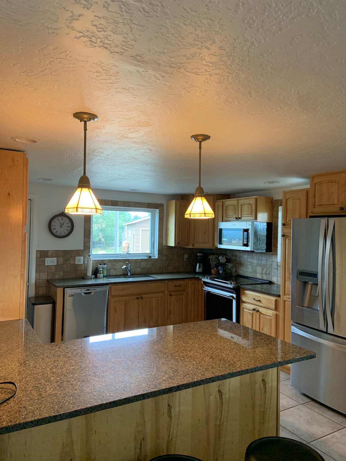 The kitchen features warm wooden cabinetry and granite countertops, highlighted by two pendant lamps. A large window provides natural light, overlooking the outdoor area. Appliances include a refrigerator, stove, dishwasher, and microwave, with a bar area equipped with seating nearby.