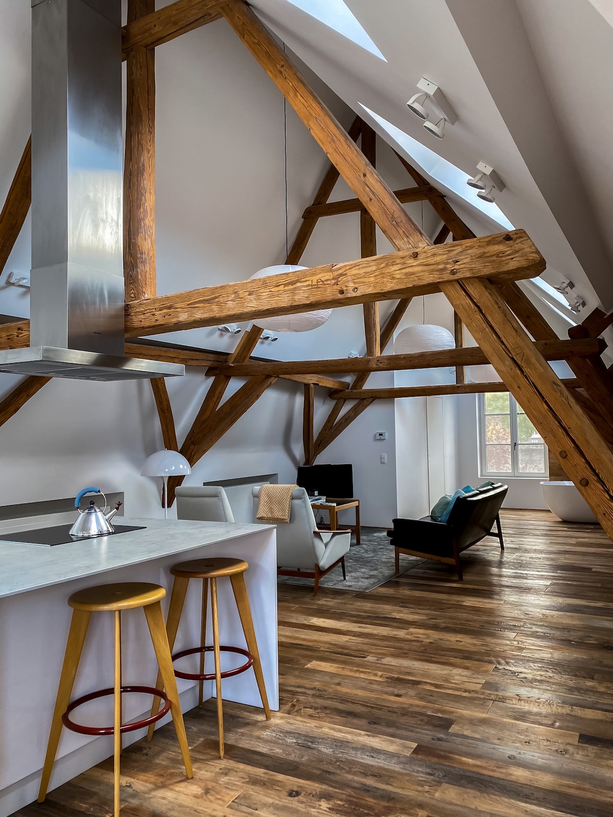 The loft features high ceilings supported by exposed wooden beams, creating an airy atmosphere. A modern kitchen is visible with sleek metal and wood accents, complemented by two high stools at the counter. Natural light enters through large windows, illuminating the stylish seating area.