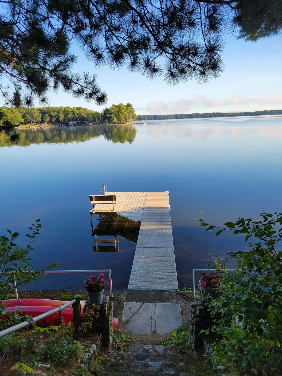 A serene lakeside scene presents a wooden dock extending into calm waters, framed by lush greenery. Kayaks rest nearby, with potted flowers adding a touch of color. The tranquil landscape reflects surrounding trees and a clear sky, inviting peaceful moments by the water.