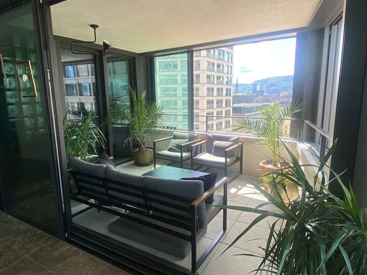 A bright balcony space features a comfortable seating arrangement with a sofa and chairs surrounded by greenery. Large windows provide panoramic views of the city, while natural light fills the area. Potted plants add a touch of nature to the inviting atmosphere.