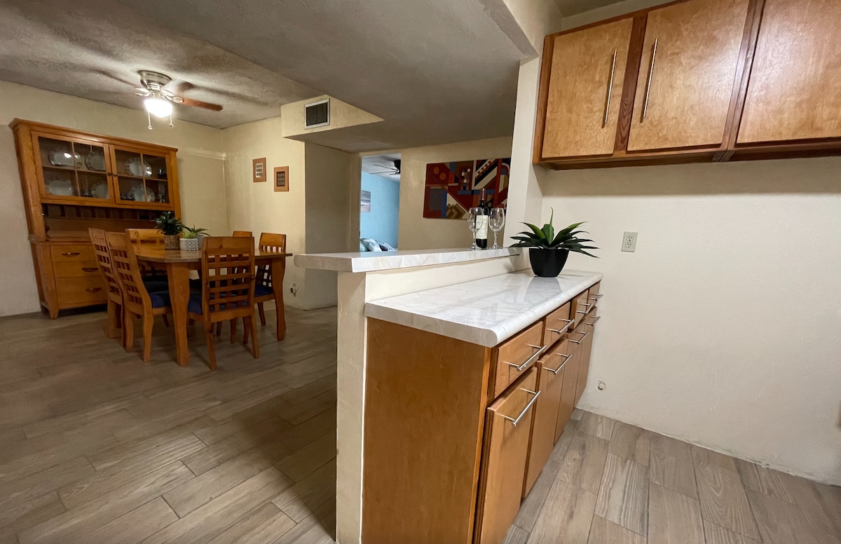 A modern kitchen area is visible, featuring light wood cabinetry and a white countertop. A dining table for six is seen in the background, complemented by a glass display cabinet. Natural light illuminates the space, enhancing the clean and organized appearance.