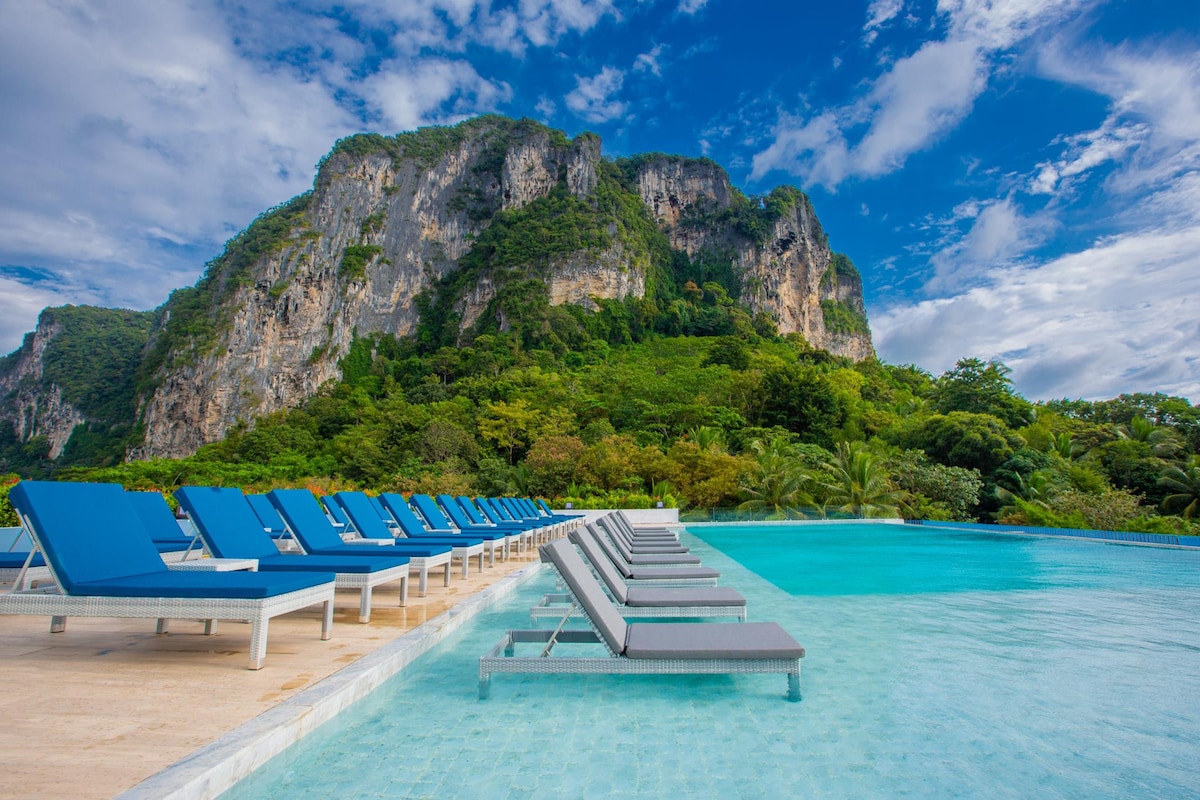 A serene outdoor pool area features multiple lounge chairs arranged neatly along the water's edge. The backdrop showcases a lush green mountain, enhancing the natural landscape. The sky is partly cloudy, with blue tones creating a calming atmosphere.