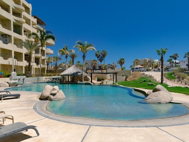 A spacious outdoor swimming pool is surrounded by palm trees and sun loungers. A thatched-roof gazebo offers shade nearby, creating an inviting space for relaxation. The clear blue water reflects the bright sky, enhancing the serene atmosphere of the common area.