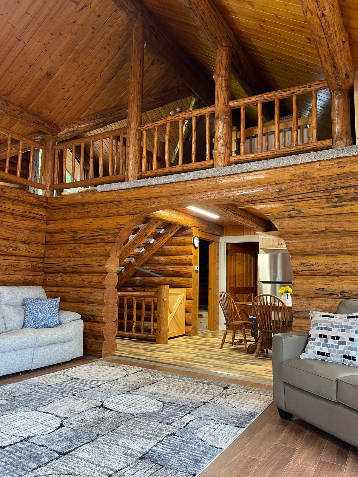 The interior of a log home showcases wooden beams and walls, highlighting natural textures. A staircase leads to an open loft area. Cozy seating arrangements including a light-colored sofa and a chair are visible, along with a dining area featuring wooden chairs in the background.