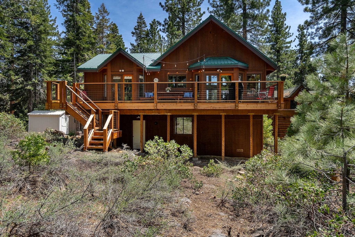 The exterior of the remodeled house features a large deck surrounded by trees, providing a serene natural setting. Stairs lead to the upper entrance, which showcases multiple windows that invite natural light. The wooden structure blends harmoniously with the lush greenery in the surrounding area.