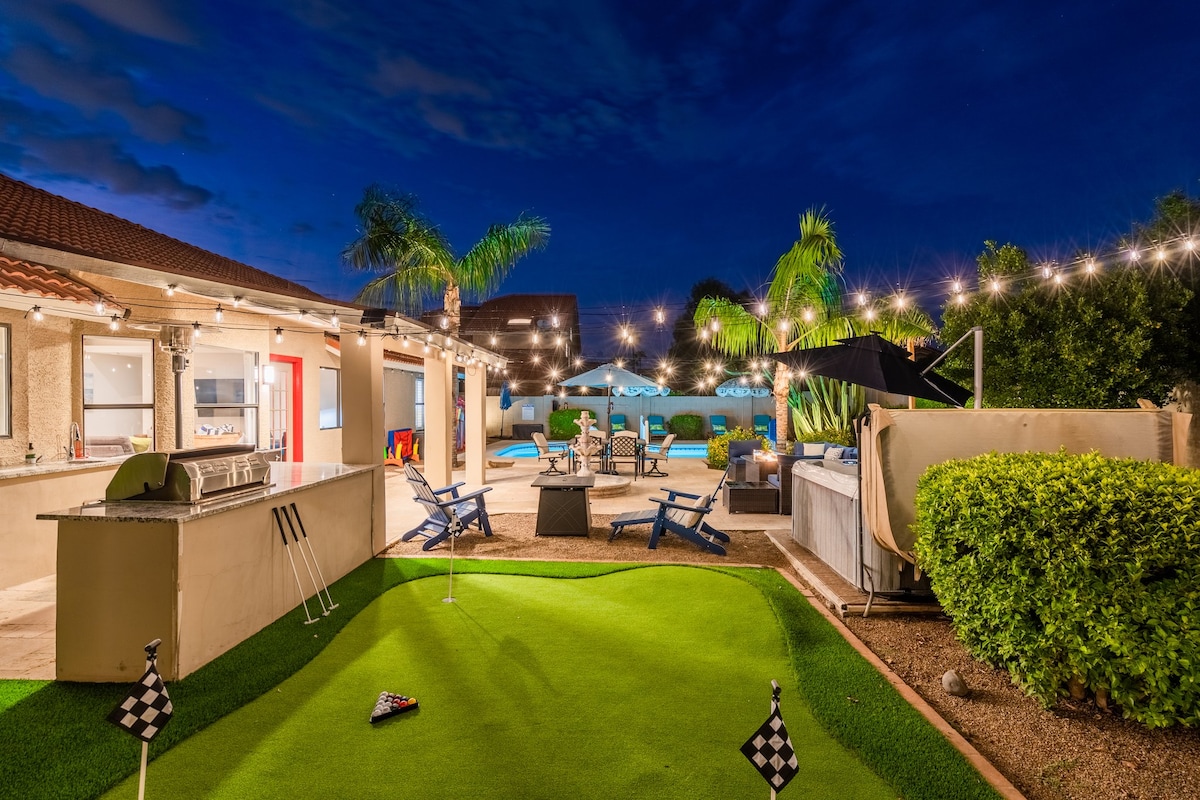 The outdoor space features a private putting green with two flags and a natural gas grill beside an outdoor kitchen. Inviting lounge areas are enhanced by string lights, and a hot tub is surrounded by palm trees and a manicured landscape, all under a night sky.
