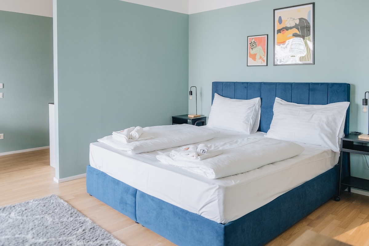 A spacious bedroom features a kingsize bed with white bedding and two pillows. The walls are painted in a soft green hue, and framed artwork hangs above the bed. Nightstands with reading lamps are placed on either side, contributing to a calming atmosphere.