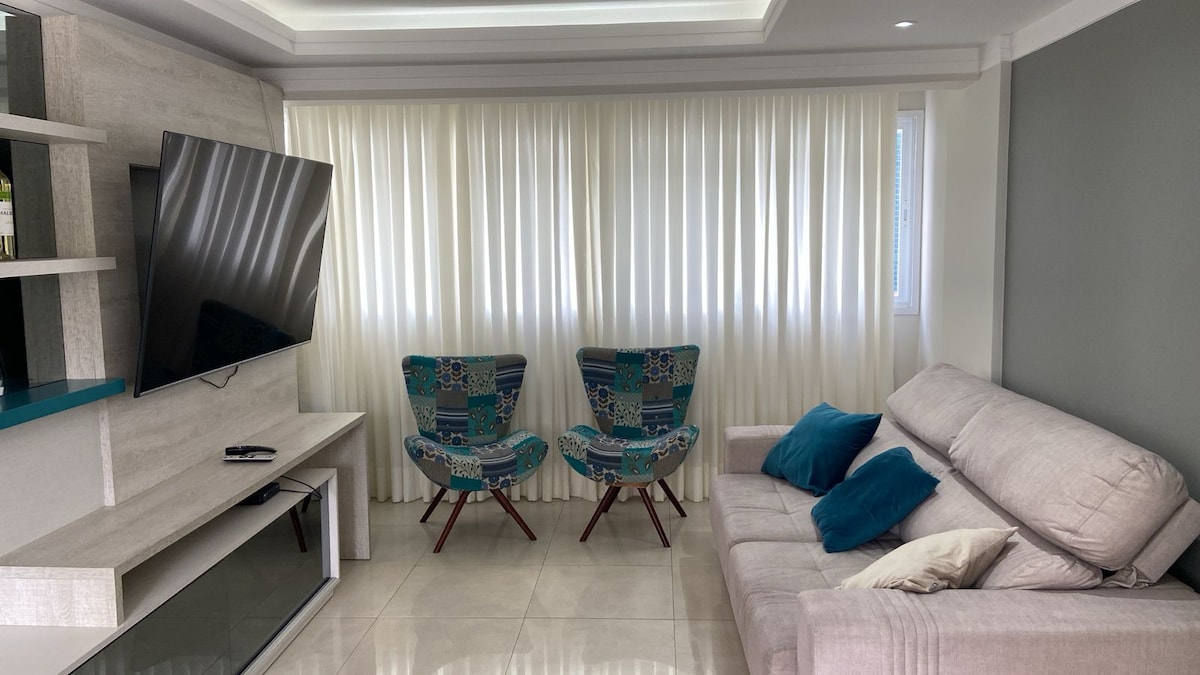 A living area is presented with a light-colored sofa, adorned with cushions. Two patterned chairs are arranged opposite a wall-mounted television. Natural light filters through the elegant vertical blinds, illuminating the neutral-toned walls and glossy tiled floor.