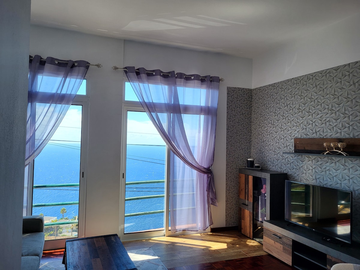 Large windows provide ample natural light and an ocean view in the living area. Stylish curtains frame the windows, while contrasting wall designs add texture. A TV stand and seating area are positioned on the wood flooring, creating a comfortable and inviting atmosphere.
