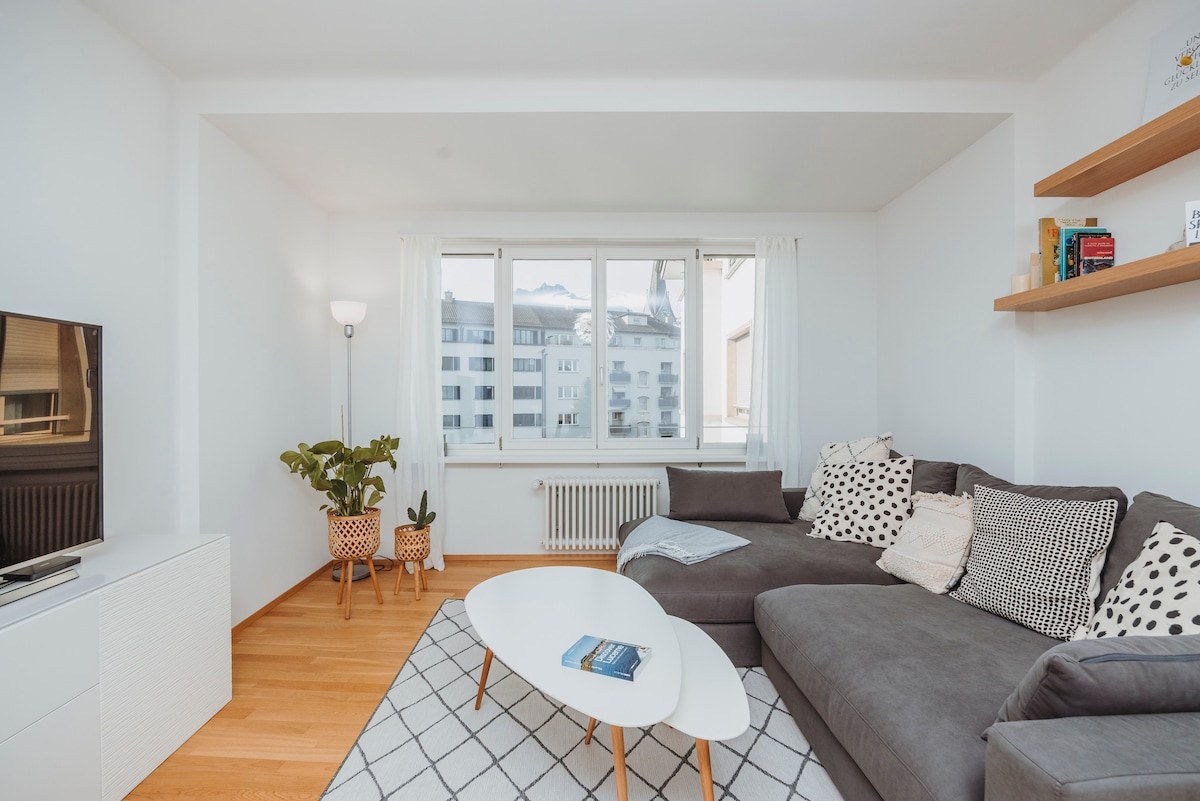 A light-filled living area features a large gray sectional sofa adorned with patterned cushions. A minimalist coffee table is positioned in front of the sofa, and a green plant adds a natural touch. Large windows overlook a scenic view, framed by soft white curtains.