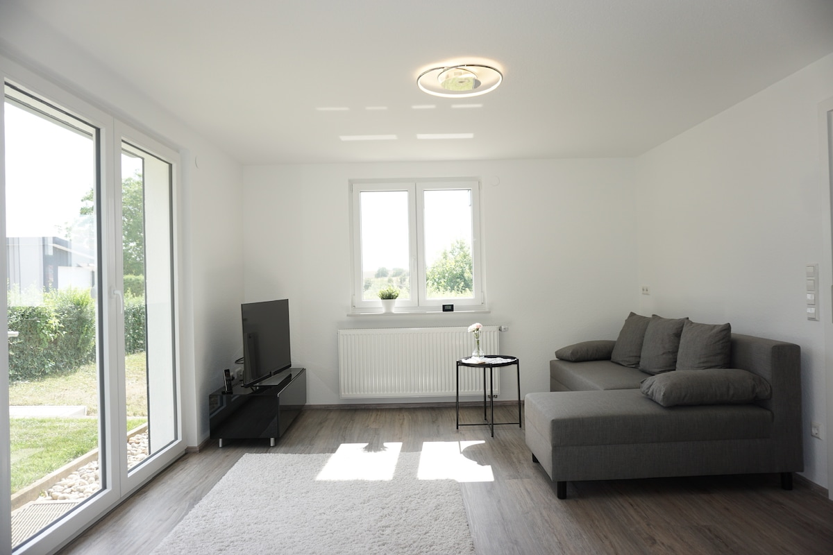 A bright living area features large windows that allow natural light to fill the space. A sectional sofa and a small table are present, along with a television mounted on the wall. Light-colored flooring complements the modern, inviting ambiance.