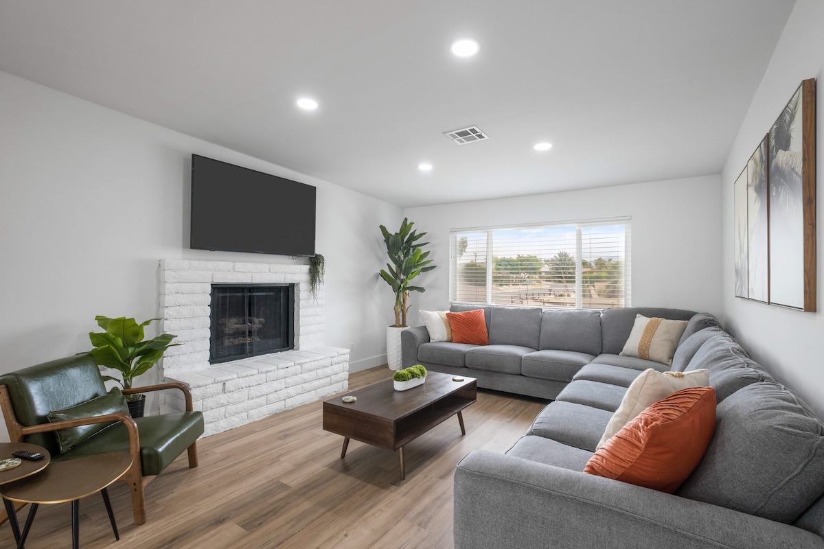 A light-filled living room features a comfortable sectional sofa with orange accent pillows. A modern coffee table sits in front of the sofa, complemented by a green armchair. A white brick fireplace serves as a focal point, with large windows providing views of the outdoors.